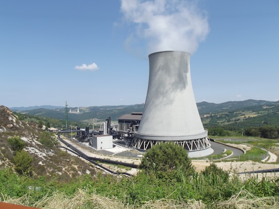 A large cooling tower is prominently visible, emitting steam against a clear blue sky. The landscape surrounding it is mountainous with patches of greenery and sparse vegetation. The power facility has several industrial structures and pipelines leading to the tower, indicating an industrial or energy generation site.