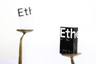A black box labeled 'Ethe' is balanced on a metallic stand in sharp focus, while another similar, white box is blurred in the background. The scene is minimalist, featuring a predominantly white backdrop, and the boxes appear to display printed details resembling packaging for a product.