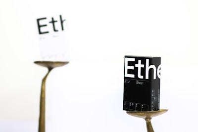 A black box labeled 'Ethe' is balanced on a metallic stand in sharp focus, while another similar, white box is blurred in the background. The scene is minimalist, featuring a predominantly white backdrop, and the boxes appear to display printed details resembling packaging for a product.