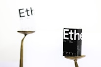 A black box labeled 'Ethe' is balanced on a metallic stand in sharp focus, while another similar, white box is blurred in the background. The scene is minimalist, featuring a predominantly white backdrop, and the boxes appear to display printed details resembling packaging for a product.