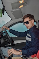 A person wearing sunglasses and a blue shirt is seated in the driver’s seat of a car, holding the steering wheel. The car interior is visible with a rearview mirror, illuminated roof lights, and a seat with a steering wheel. The person is wearing a wristwatch and seems casual and confident.