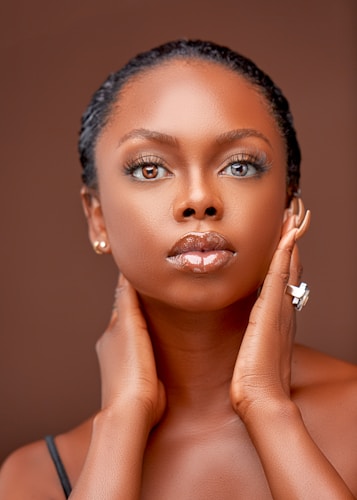 A close-up portrait of a person with smooth skin and glossy lips, gazing directly at the camera. The person has their hands gently placed on their neck, showcasing a sense of confidence and poise. The background is a warm, muted brown that complements the subject's skin tone.