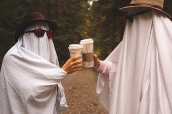 Two individuals dressed as playful ghosts, wearing sunglasses and hats, clink disposable coffee cups together. They are standing on a forest path surrounded by trees, creating a whimsical and humorous atmosphere.