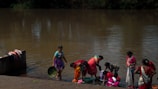 A group of women is gathered by a riverbank, engaging in laundry activities. They are wearing colorful traditional garments and using the water from the river to wash clothes. Nearby, clothes are draped over a wall to dry. The brown water reflects the surrounding greenery.