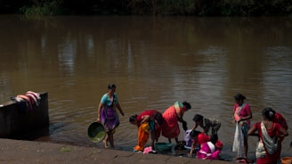 A group of women is gathered by a riverbank, engaging in laundry activities. They are wearing colorful traditional garments and using the water from the river to wash clothes. Nearby, clothes are draped over a wall to dry. The brown water reflects the surrounding greenery.