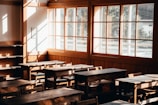 A rustic classroom setting with wooden desks and chairs is illuminated by soft, warm sunlight streaming through large windows. The sunlight casts gentle shadows across the room, emphasizing the natural wood tones of the furniture and walls.