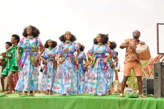 A group of women in colorful, traditional dresses perform a dance on a green platform. A man beside them plays a drum, wearing a light brown outfit. Other performers in vibrant clothing stand in the background.