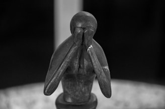 A wooden sculpture depicts a seated figure with elongated limbs, head resting on hands in a pose conveying thoughtfulness or sadness. The abstract design focuses on simple lines and a smooth texture, highlighted in shades of black and gray.
