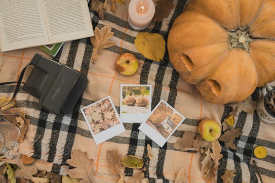 A top-down view of an autumn-themed arrangement featuring a plaid blanket on which lies a large pumpkin, dried leaves, and a few apples. There's an open book, a lit candle, and a vintage Polaroid camera with three instant photos placed nearby. The overall setup conveys a cozy, seasonal atmosphere.