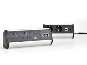 Two multi-functional power strips with various power and data ports are placed on a white surface. The power strips feature UK and European sockets, USB ports, and Ethernet connections. They have a modern design with metallic and black elements.