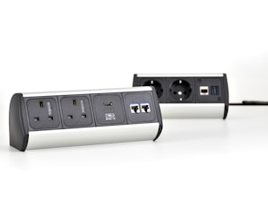 Two multi-functional power strips with various power and data ports are placed on a white surface. The power strips feature UK and European sockets, USB ports, and Ethernet connections. They have a modern design with metallic and black elements.
