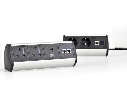 Two multi-functional power strips with various power and data ports are placed on a white surface. The power strips feature UK and European sockets, USB ports, and Ethernet connections. They have a modern design with metallic and black elements.