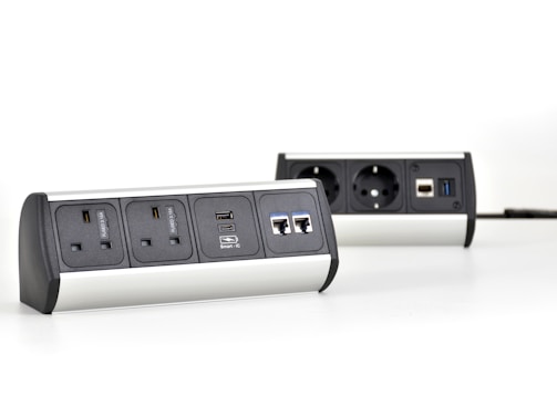 Two multi-functional power strips with various power and data ports are placed on a white surface. The power strips feature UK and European sockets, USB ports, and Ethernet connections. They have a modern design with metallic and black elements.