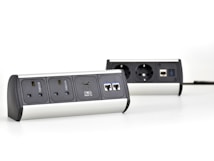 Two multi-functional power strips with various power and data ports are placed on a white surface. The power strips feature UK and European sockets, USB ports, and Ethernet connections. They have a modern design with metallic and black elements.