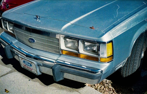 A vintage blue Ford car with a prominent grille and headlights. The car's surface shows some wear with a leaf resting on the hood. The front license plate reads 'Ford Racing.' The tires are visibly aged, and there are fallen leaves on the ground.