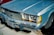 A vintage blue Ford car with a prominent grille and headlights. The car's surface shows some wear with a leaf resting on the hood. The front license plate reads 'Ford Racing.' The tires are visibly aged, and there are fallen leaves on the ground.