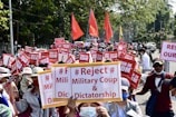 A large group of people is gathered in a protest on a city street, holding signs with messages rejecting a military coup and dictatorship. Red flags and banners are visible among the crowd, and many participants are wearing hats and face masks. The surroundings include trees and vehicles parked on the side of the road.