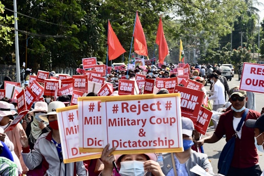 A large group of people is gathered in a protest on a city street, holding signs with messages rejecting a military coup and dictatorship. Red flags and banners are visible among the crowd, and many participants are wearing hats and face masks. The surroundings include trees and vehicles parked on the side of the road.