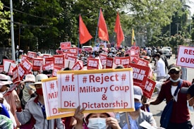 A large group of people is gathered in a protest on a city street, holding signs with messages rejecting a military coup and dictatorship. Red flags and banners are visible among the crowd, and many participants are wearing hats and face masks. The surroundings include trees and vehicles parked on the side of the road.