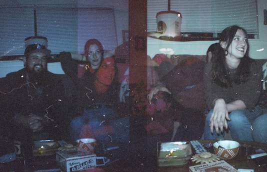 A dimly lit room features a group of people sitting together. The lighting creates a moody, nostalgic effect with visible film grain and a slight red and blue tone overlay. Various objects, including snack containers and a pizza box, are on the table in front of them. The individuals appear to be casually dressed, engaged in conversation, or laughter.