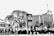 Black and white photo depicting a blurred crowd walking past a large building with geometric patterns and trees. The scene suggests motion, possibly a march or protest, with urban architecture and overhead power lines.