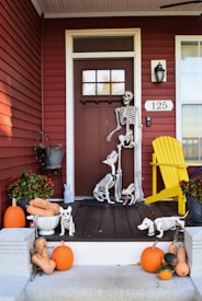 A porch decorated for Halloween with skeleton figures of a human and various animals, such as dogs and a cat. Several pumpkins and gourds are arranged around the steps, alongside a mix of potted autumn plants. The porch features a red door with the number 125, an outdoor lantern, and a yellow chair.