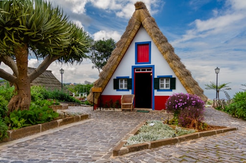 A charming, small A-frame house features a thatched roof and a red, blue, and white color scheme. The house is surrounded by vibrant greenery and colorful flowers, with a cobblestone pathway leading to the entrance. A large, distinctive tree is to the left, and there are stone planters filled with lush plants.