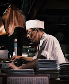An elderly man wearing a white chef's hat and uniform is focused on cooking. He appears to be in a dimly lit kitchen, surrounded by various cooking tools and stacks of plates. A large piece of dried fish hangs overhead.