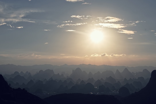 The image captures a serene landscape with rolling hills silhouetted against a bright sun, casting gentle rays and shadows on the horizon. Wispy clouds are spread across a clear sky, adding depth and contrast to the scene.
