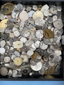A collection of various vintage watch faces and mechanical watch movements, with intricate gears and dials displayed in a box. The assortment displays a range of colors from metallic silver and brass to old, worn-out tones. Some watch faces have visible roman numerals or numbers, and the components are stacked and scattered in no particular order.