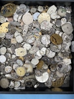 A collection of various vintage watch faces and mechanical watch movements, with intricate gears and dials displayed in a box. The assortment displays a range of colors from metallic silver and brass to old, worn-out tones. Some watch faces have visible roman numerals or numbers, and the components are stacked and scattered in no particular order.
