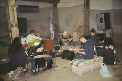 A group of musicians are gathered in a rehearsal space. There are five people with various musical instruments, including a drum set, keyboard, and guitars. The room seems industrial, with exposed beams and scattered equipment. The atmosphere is casual, with some musicians seated on makeshift seating like pallets and cushions.