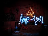 A room with a wall covered in sketches and illustrations, lit by small, star-like lights arranged in a triangular shape. In front is a bed with a colorful quilt. Glowing blue light trails spell out 'love you' in playful cursive against a dark background.