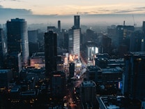 A bustling cityscape at dusk featuring tall modern skyscrapers illuminated with lights. The streets are busy, and large digital billboards display vibrant advertisements. The sky is a mix of cool blues and warm tones as the sun sets.