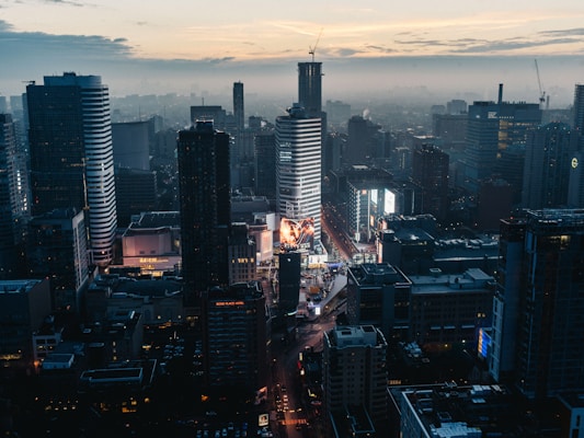A bustling cityscape at dusk featuring tall modern skyscrapers illuminated with lights. The streets are busy, and large digital billboards display vibrant advertisements. The sky is a mix of cool blues and warm tones as the sun sets.