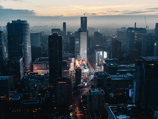 A bustling cityscape at dusk featuring tall modern skyscrapers illuminated with lights. The streets are busy, and large digital billboards display vibrant advertisements. The sky is a mix of cool blues and warm tones as the sun sets.