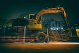 A construction site under dim lighting, featuring a large excavator behind a meshed fence. The scene is set in an urban area with tall buildings in the background, illuminated by artificial lights.