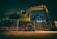 A construction site under dim lighting, featuring a large excavator behind a meshed fence. The scene is set in an urban area with tall buildings in the background, illuminated by artificial lights.