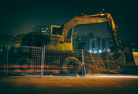 A construction site under dim lighting, featuring a large excavator behind a meshed fence. The scene is set in an urban area with tall buildings in the background, illuminated by artificial lights.