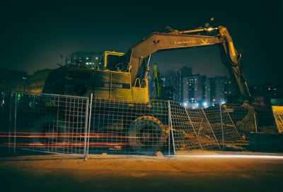 A construction site under dim lighting, featuring a large excavator behind a meshed fence. The scene is set in an urban area with tall buildings in the background, illuminated by artificial lights.