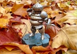 A figure of a medieval knight in silver armor stands on a grey pedestal, surrounded by a layer of autumn leaves in shades of orange, red, and yellow. The armored figure has a helmet with a visor and holds a small shield.