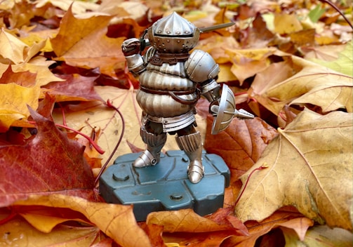 A figure of a medieval knight in silver armor stands on a grey pedestal, surrounded by a layer of autumn leaves in shades of orange, red, and yellow. The armored figure has a helmet with a visor and holds a small shield.