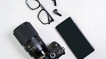 A flat lay of technology items including a camera with a large lens, eyeglasses, wireless earbuds, and a smartphone placed on a white surface.