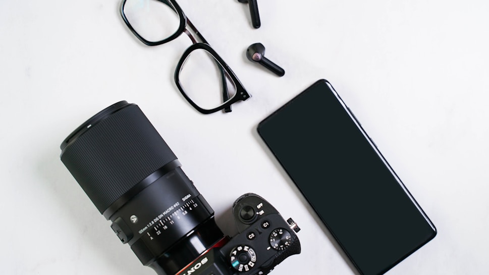 A flat lay of technology items including a camera with a large lens, eyeglasses, wireless earbuds, and a smartphone placed on a white surface.