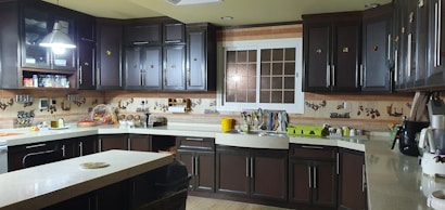 A kitchen with dark wood cabinetry and beige countertops, illuminated by overhead lighting. Several kitchen appliances, including a stove, a blender, and a microwave, are visible. Various kitchen utensils hang from the wall, and a window is positioned above the sink. There are dishes drying on a rack near the sink.