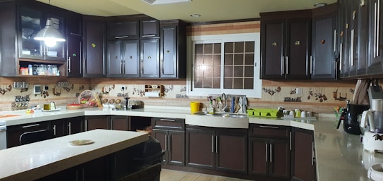 A kitchen with dark wood cabinetry and beige countertops, illuminated by overhead lighting. Several kitchen appliances, including a stove, a blender, and a microwave, are visible. Various kitchen utensils hang from the wall, and a window is positioned above the sink. There are dishes drying on a rack near the sink.
