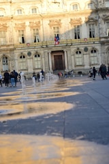 A grand stone building with ornate architecture stands in the background, flanked by large windows and decorative details. In the foreground, water fountains create a playful pattern on the paved ground. People are casually walking and interacting, some with children, enjoying the public space as the golden light casts reflections on the wet pavement.