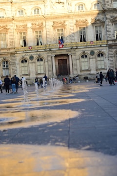 A grand stone building with ornate architecture stands in the background, flanked by large windows and decorative details. In the foreground, water fountains create a playful pattern on the paved ground. People are casually walking and interacting, some with children, enjoying the public space as the golden light casts reflections on the wet pavement.