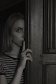 A person stands partially obscured by a wooden door, holding a finger to their lips in a gesture suggesting silence or secrecy. The lighting is dim, creating a mysterious and slightly ominous atmosphere.