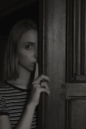 A close-up of a mysterious figure whispering in a dimly lit room filled with shadows.